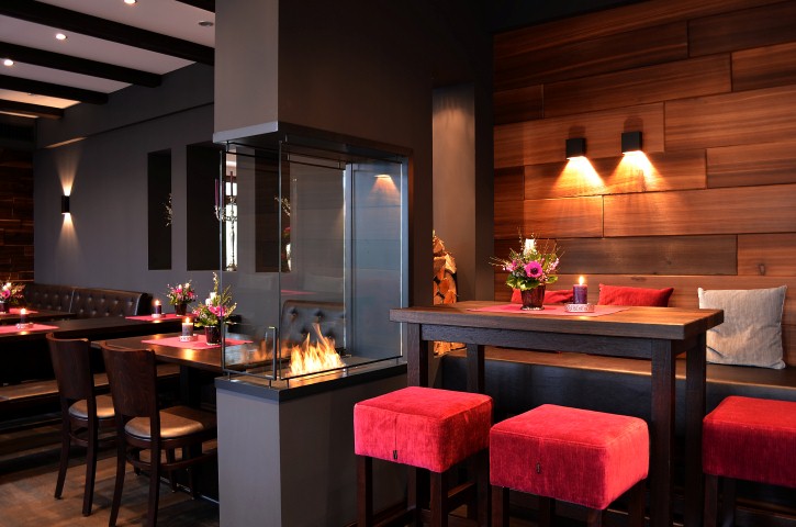 Modernes Restaurantinterieur mit einem gläsernen Kamin, roten Hockern, Holztischen und Blumengestecken – ein idealer Veranstaltungsort für Ihre nächste private Feier oder eine unvergessliche Hochzeitslocation.