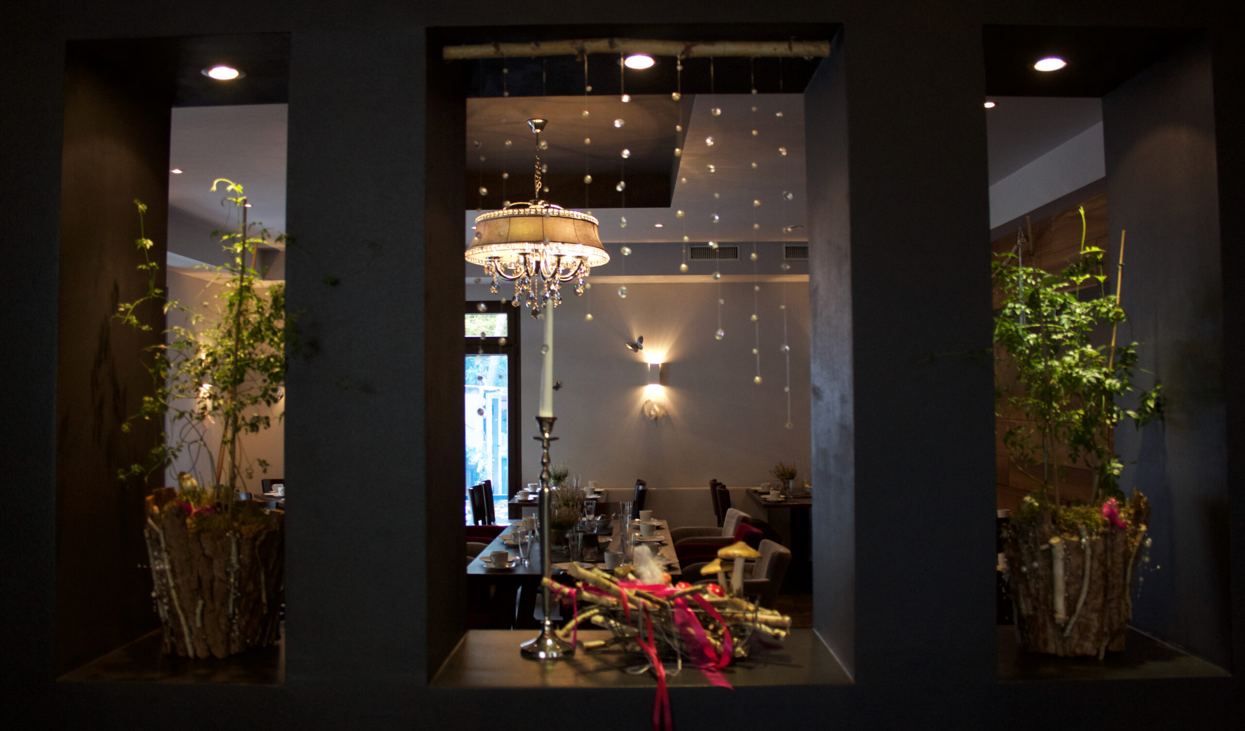 Ein elegantes Restaurantinterieur, das durch drei dunkel gerahmte Fenster zu sehen ist, mit Pflanzen und einem Kronleuchter im Inneren – ideal als Hochzeitslocation oder für eine unvergessliche Privatfeier, mit der Möglichkeit einer Übernachtung für die Gäste.