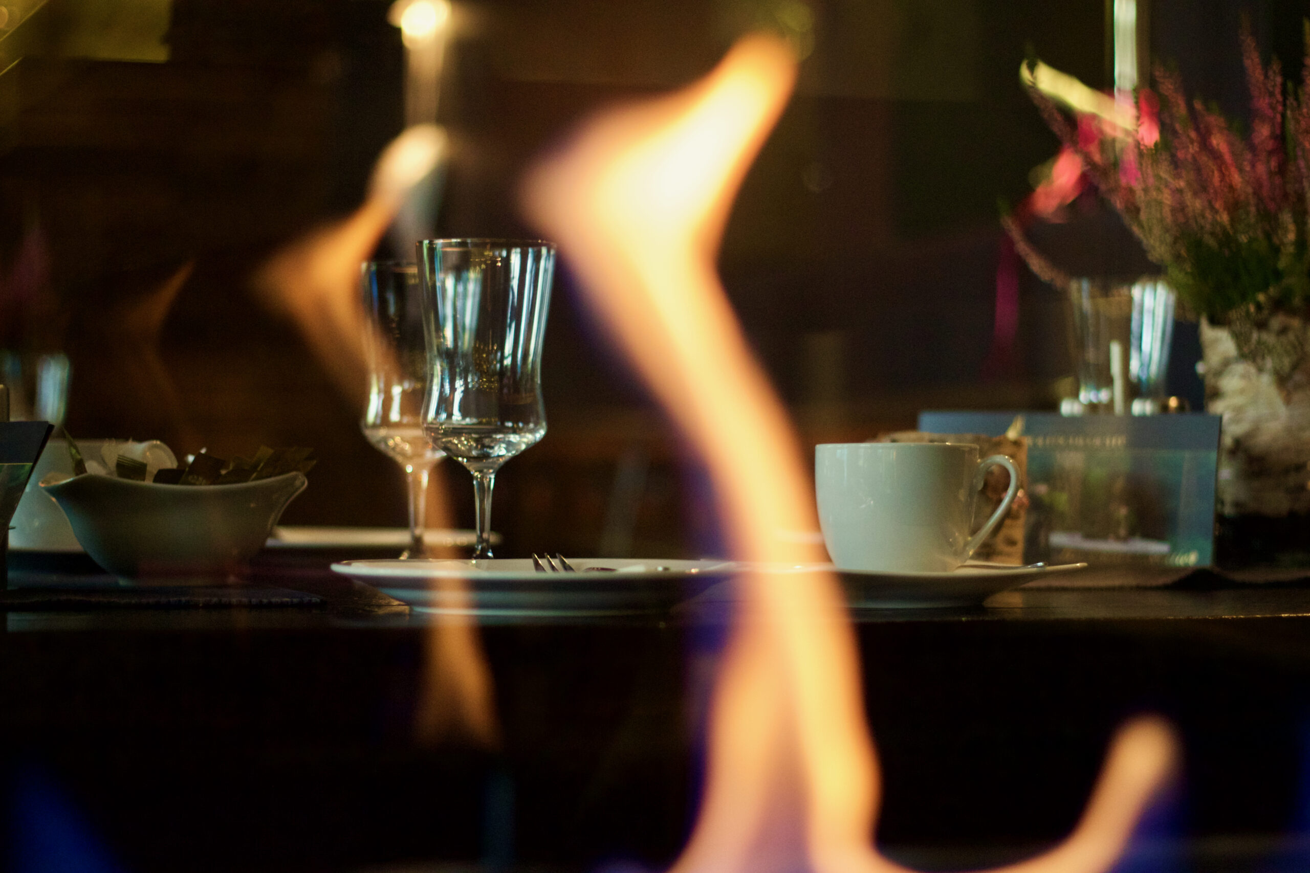 Ein mit Gläsern und einer Tasse gedeckter Tisch, der durch die Flammen eines Feuers im Vordergrund zu sehen ist, schafft eine intime Atmosphäre – perfekt für eine Privatfeier oder als elegante Hochzeitslocation.
