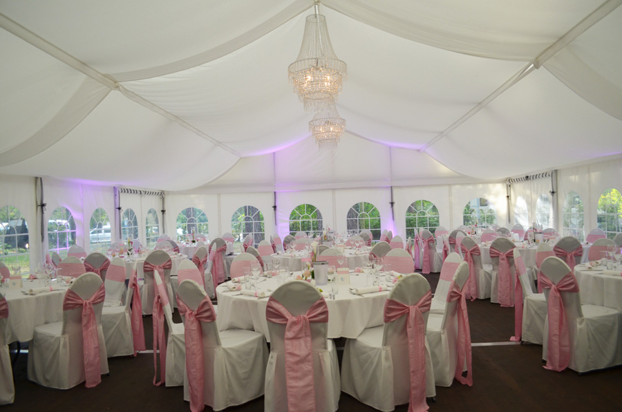 Ein weißes Zelt mit runden Tischen, weißen Stühlen mit rosa Schleifen und Kronleuchtern, die von der Decke hängen – ein eleganter Veranstaltungsort, perfekt für Ihre unvergessliche Feier.