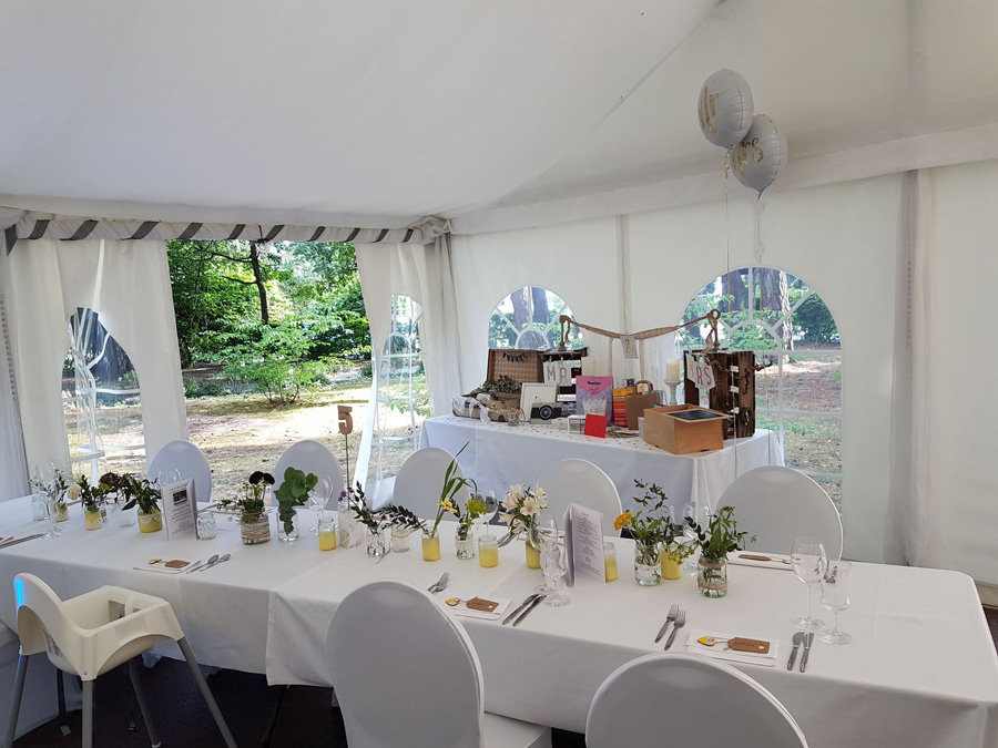 Ein weißes Zelt mit runden Tischen, die für ein Essen gedeckt sind, im Hintergrund ein mit Blumen und Geschenken geschmückter Tisch – perfekt als elegante Hochzeitslocation oder für eine unvergessliche Privatfeier.