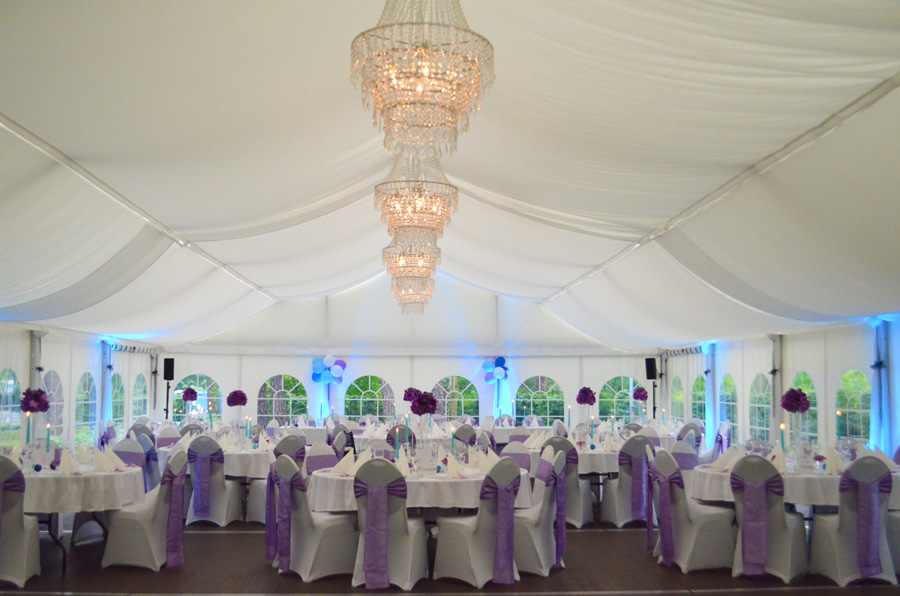 Eine elegante Veranstaltungslocation mit runden Tischen, Kronleuchtern und violetten Stuhlschleifen unter einem weißen Festzelt – perfekt für Ihre nächste Firmenfeier oder ein großes Fest.