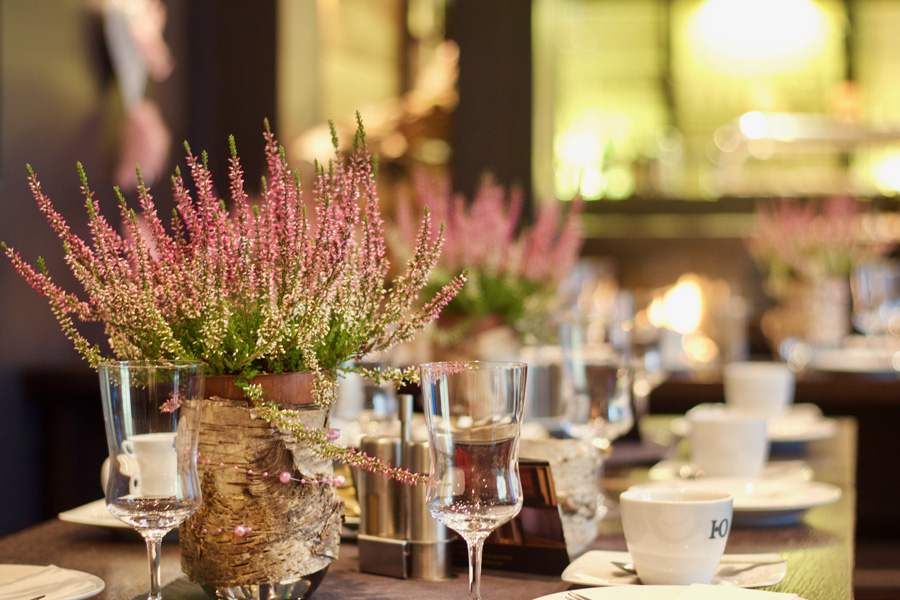 Rosa Blumen in Vasen schmücken einen Tisch, der mit Gläsern, Tassen und Tellern gedeckt ist, in einem sanft beleuchteten Raum und schaffen so die perfekte Atmosphäre für eine elegante Hochzeitslocation oder eine unvergessliche Firmenfeier.
