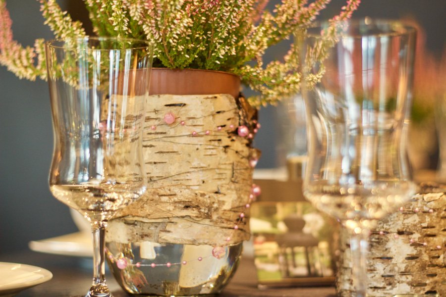 Auf einem sanft unscharf abgebildeten Tisch stehen klare Weingläser und eine mit Birkenrinde umwickelte Vase mit rosa Blumen, die den eleganten Charme einer Hochzeitslocation vermitteln, die sich perfekt für ein unvergessliches Fest eignet.