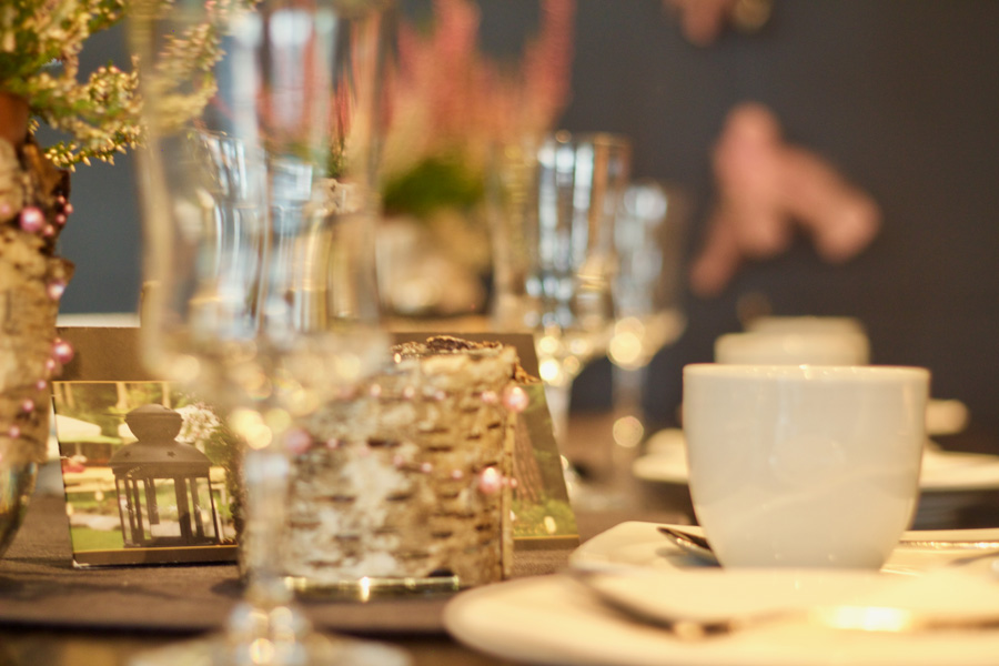 Eine weiße Kaffeetasse mit Untertasse auf einem für eine Mahlzeit gedeckten Tisch, im Hintergrund verschwommene Gläser und Dekorationen – perfekt für eine elegante Veranstaltungs- oder Hochzeitslocation, an der die Gäste jedes Detail genießen und sich nach der Feier sogar bei einer Übernachtung entspannen können.