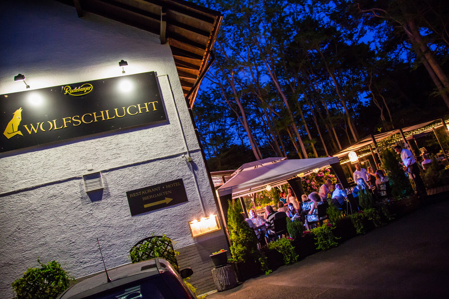 Nachtansicht eines Restaurants im Freien, in dem Gäste unter Lichterketten speisen, und ein Schild mit der Aufschrift „Wolfsschlucht“ – damit ist es der ideale Ort für Hochzeiten oder Veranstaltungen wie eine unvergessliche Firmenfeier.