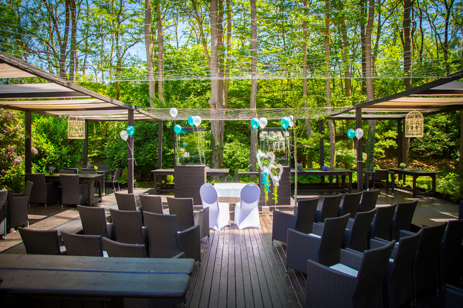 Eine Hochzeitskulisse im Freien mit schwarzen Stühlen, blauen und weißen Luftballons, einer Holzterrasse und Bäumen im Hintergrund – diese wunderschöne Hochzeitslocation ist perfekt für unvergessliche Feierlichkeiten.