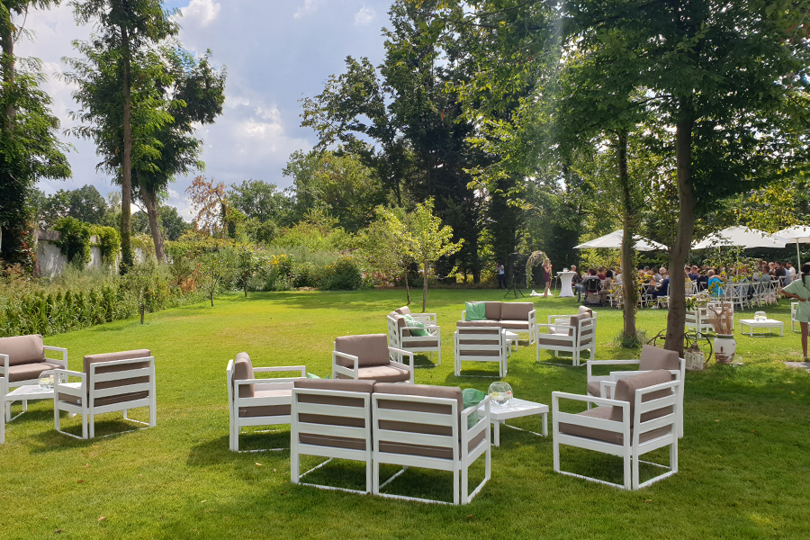 Auf dem grünen Rasen stehen weiße Gartenstühle und -tische, umgeben von Bäumen und Menschen, die sich unter Sonnenschirmen versammelt haben – ein idealer Veranstaltungsort für eine Firmenfeier oder eine Privatfeier.