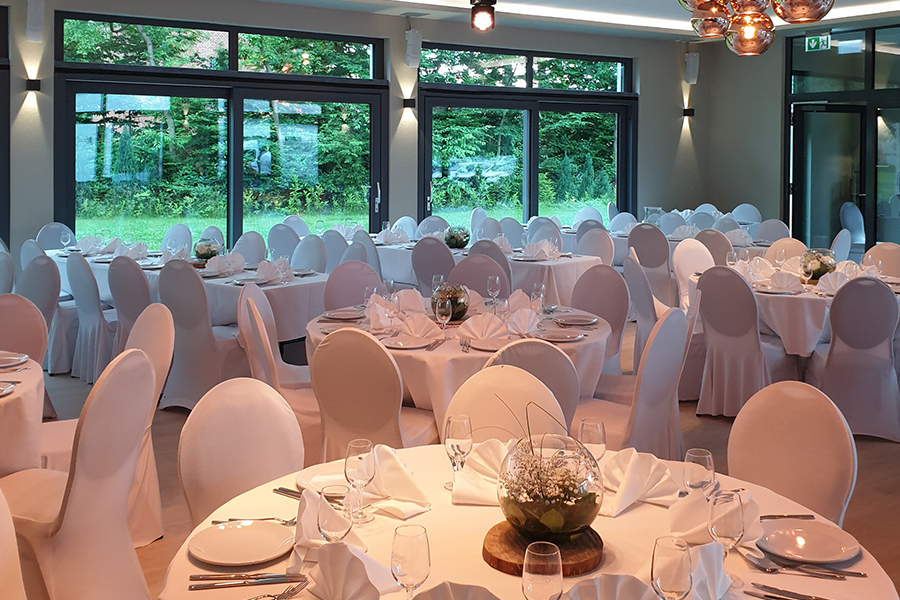 Ein eleganter Festsaal mit runden Tischen, weißer Tischwäsche und großen Fenstern mit Blick ins Grüne – ideal als Hochzeitslocation oder für eine unvergessliche Privatfeier.