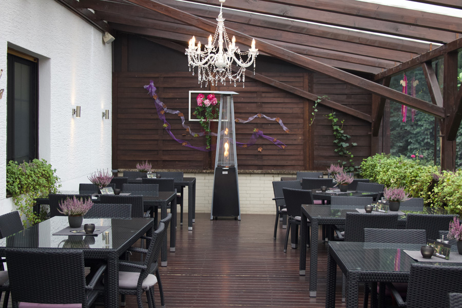 Eine Terrasse im Freien mit schwarzen Korbmöbeln, einem Kronleuchter und violetten Blumendekorationen an der Wand – eine einladende Hochzeitslocation, die sich gleichermaßen perfekt für eine entspannte Übernachtung wie auch für eine elegante Firmenfeier eignet.