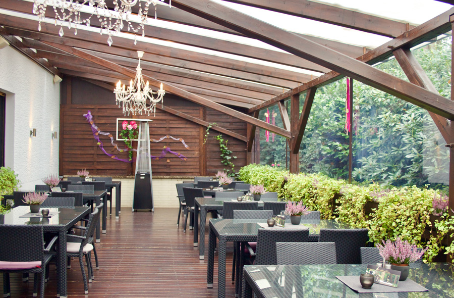 Eine Terrasse im Freien mit Glasdach, schwarzen Tischen, lila Blumen und einem Kronleuchter, umgeben von viel Grün – der ideale Rahmen für Ihre Privat- oder Firmenfeier mit der Möglichkeit zur Übernachtung.